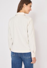 Camicia color crema con colletto ripiegato, maniche lunghe con dettagli di bottoni e una vestibilità comoda, abbinata a jeans blu.
