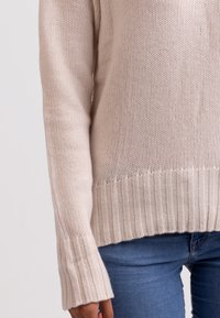 Beige Strickpullover mit strukturiertem Design, geripptem Saum und Bündchen sowie lockerer Passform. Getragen mit blauen Jeans.