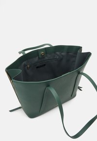 Grøn lærred-taske med et rummeligt indre, der har en mørk foring og en lynlåslomme. Lange håndtag og en lukkedetalje med trykknap.