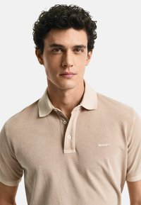 Beige polo skjorta med strukturstickning, korta ärmar och krage, med en subtil logotyp på vänster sida.