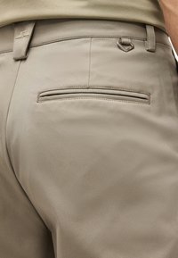 Κοντινό πλάνο μπεζ παντελονιού με πίσω εξωτερική τσέπη και ένα μικρό ασημένιο D-δακτύλιο προσαρτημένο κοντά στη μέση.