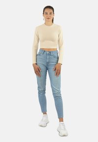 Beige Langarm-Crop-Top und hellblaue hochtaillierte Jeans. Weiße Sneakers. Das Model steht mit den Händen an den Seiten vor einem schlichten Hintergrund.