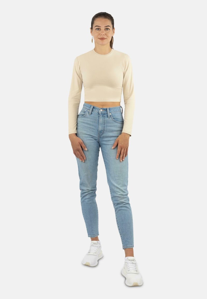 Beige Langarm-Crop-Top und hellblaue hochtaillierte Jeans. Weiße Sneakers. Das Model steht mit den Händen an den Seiten vor einem schlichten Hintergrund.