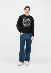 Μαύρο φούτερ με γραφικό τύπωμα και κείμενο "SERVICE WORK", σε συνδυασμό με φαρδιά μπλε τζιν παντελόνια και ανοιχτόχρωμα αθλητικά παπούτσια.