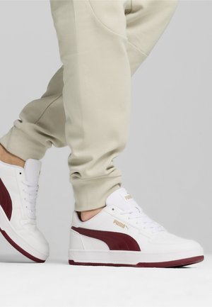 Sneaker bianche con una striscia laterale bordeaux, materiale in pelle liscia, suola in gomma piatta e marchio dorato sul lato. Indossate con pantaloni jogger beige.