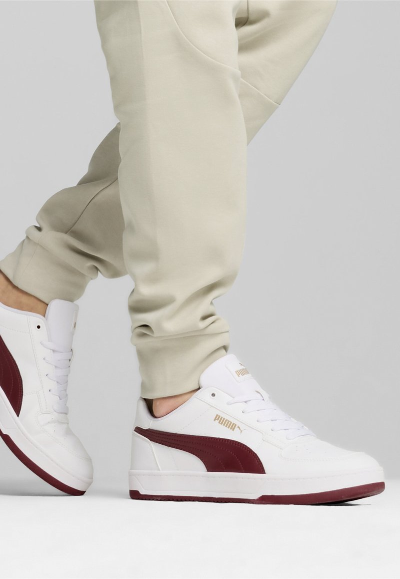 Valged tennisetossud burgundia punase küljeribaga, sileda nahkmaterjaliga, lameda kummitald ja kuldne brändimine küljel. Kannab beeže joggers'eid.