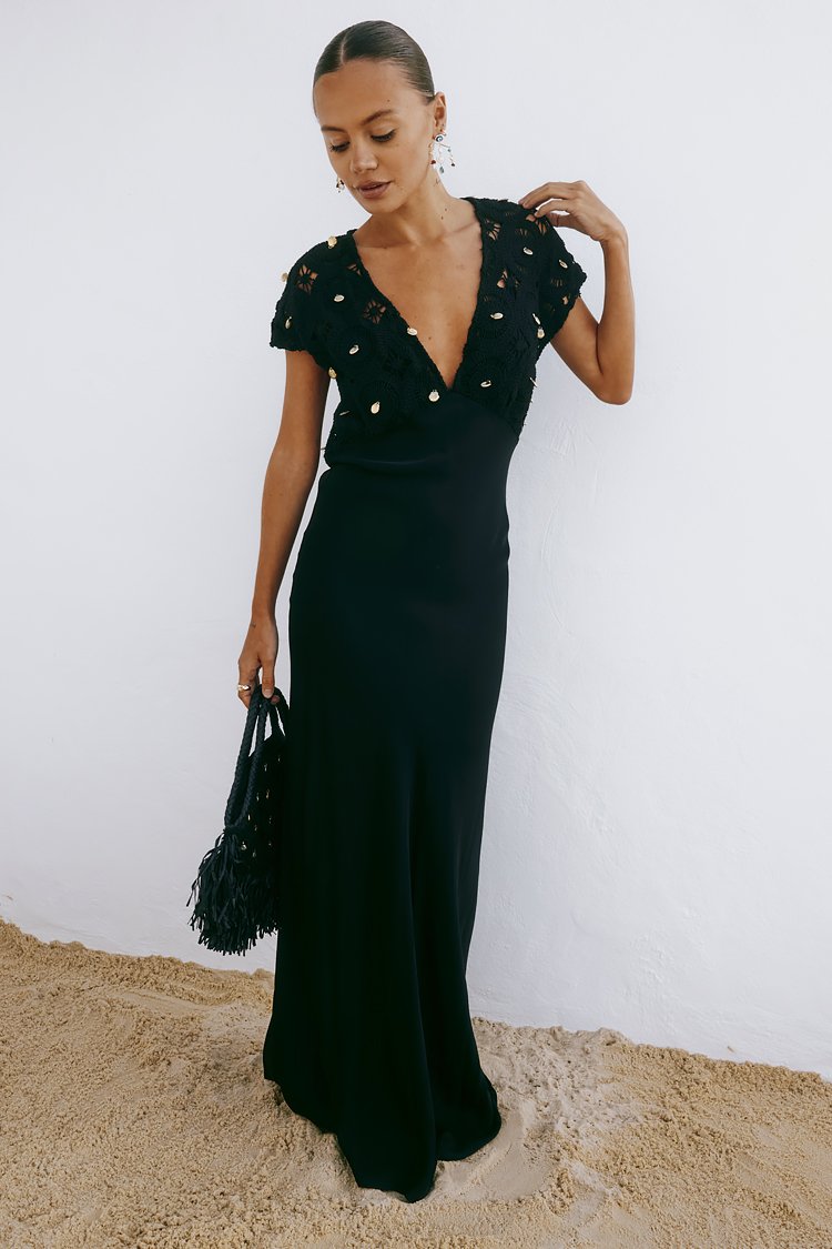 Schwarzes Maxikleid mit kurzen Spitzensleeves und tiefem V-Ausschnitt. Besitzt eine figurbetonte Silhouette und eine glatte Textur. Ergänzt durch eine strukturierte schwarze Tasche.