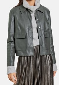 Veste en cuir vert foncé avec un col, deux poches avant et des fermetures à pression, superposée à un pull en maille gris et une jupe plissée métallique.