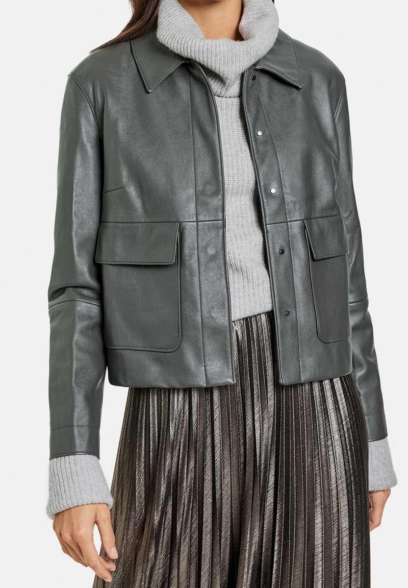 Veste en cuir vert foncé avec un col, deux poches avant et des fermetures à pression, superposée à un pull en maille gris et une jupe plissée métallique.