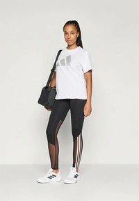 Biele tričko s šedým logom, čierne legíny s sieťovanými panelmi a tromi bielymi pruhmi. Čierna taška na rameno a biele tenisky.