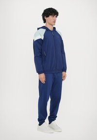 Navy windbreaker with light blue accents, zip closure, and two zippered pockets, paired with matching navy joggers and white sneakers.