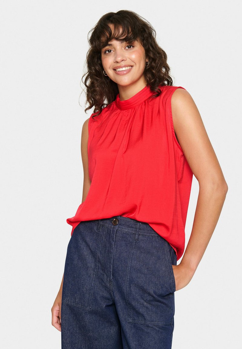 Top rosso senza maniche con collo alto, caratterizzato da dettagli arricciati e una texture liscia, abbinato a pantaloni in denim scuro.