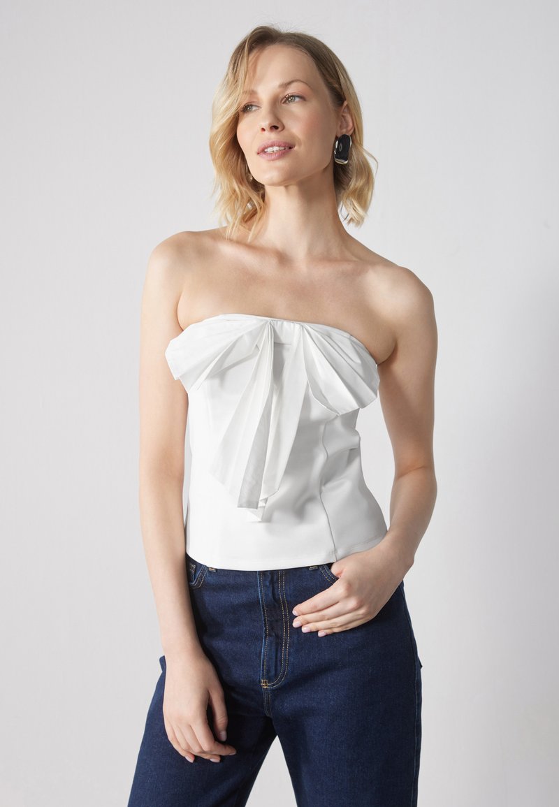 Hvit stroppeløs topp med plisserte detaljer på forsiden, tettsittende silhuett, glatt stoff. Kombinert med mørke denimjeans som viser en uformell stil.