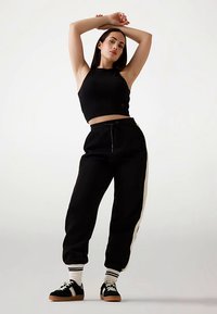 Czarny krótki top i czarne joggery z białymi paskami po bokach, zestawione z czarnymi tenisówkami i białymi skarpetkami w paski, na jednolitym tle.