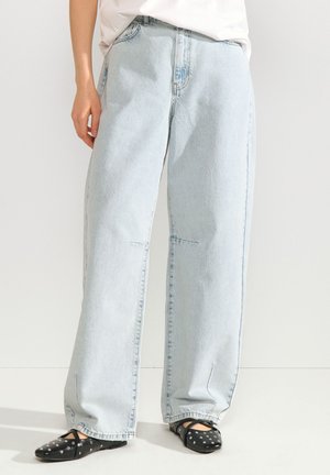 Personne portant un jean bleu clair ample et des ballerines noires à pois blancs, debout devant un fond clair uni.