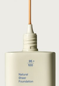 Fond de teint transparent naturel dans un flacon en crème avec une pompe, liquide beige clair s'écoulant du bout, texte bleu avec les détails du produit.