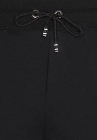 Ceinture en tissu noir avec un cordon de serrage noir passé à travers des œillets métalliques, extrémités ornées de bouts en métal argenté.