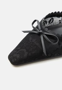 Chaussure plate noire en dentelle à bout pointu avec des bords en forme de coquille et un petit nœud sur le dessus, ornée de détails à motif floral.