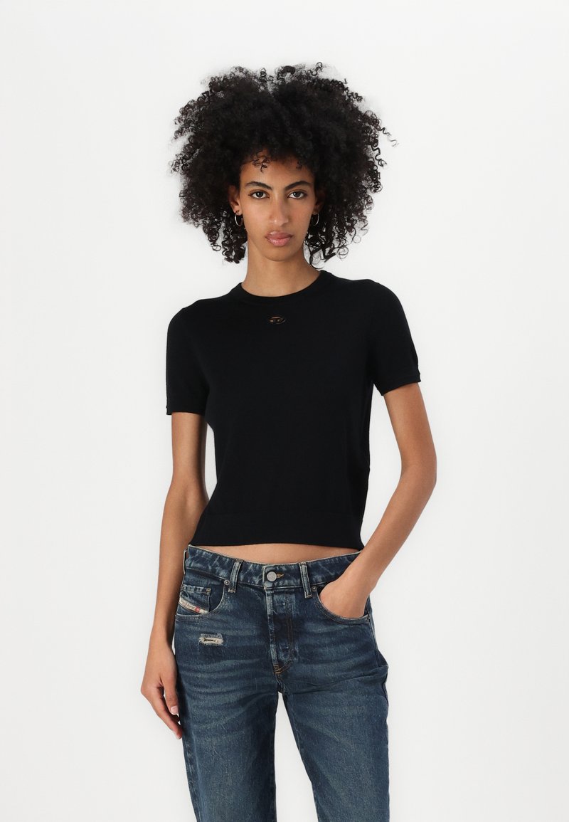 T-shirt cropped in cotone nero con maniche corte e scollatura tonda, caratterizzato da un piccolo logo metallico. Abbinato a jeans blu strappati.