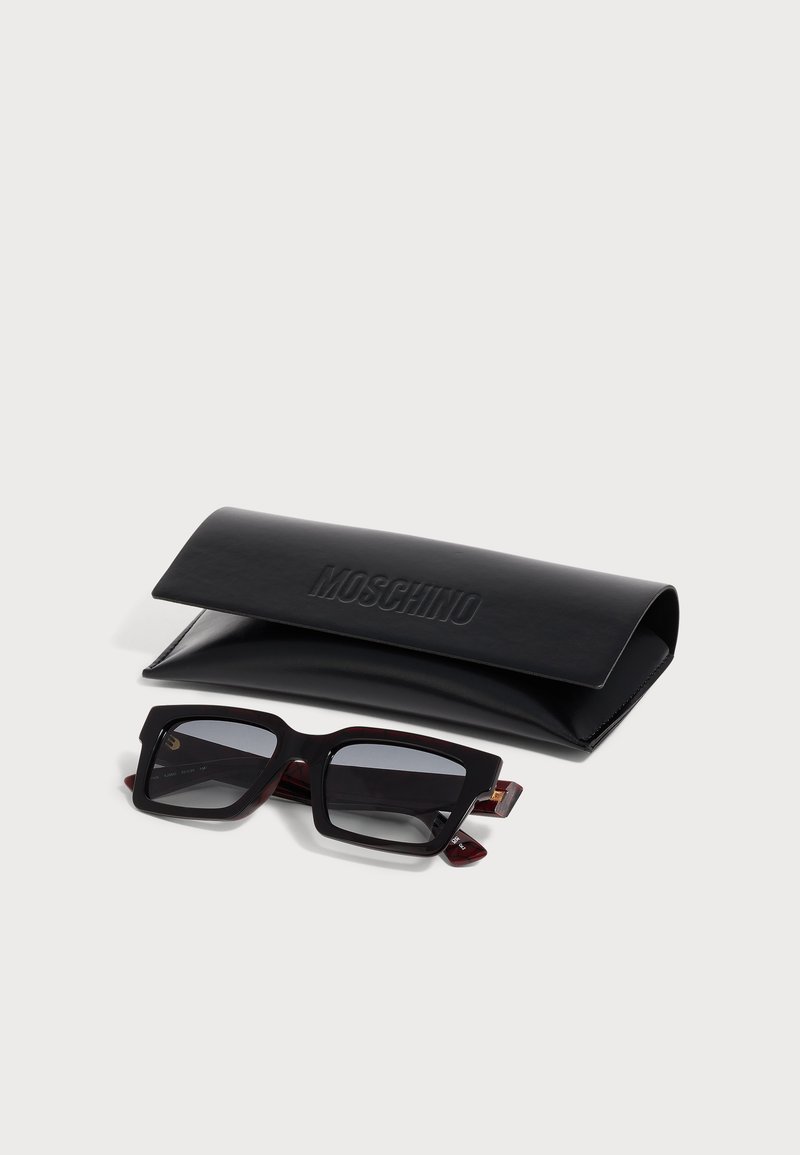 Black rectangular sunglasses with dark lenses resting in front of a black Moschino branded leather case on a white background.