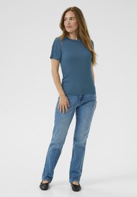 Türkises Kurzarmoberteil mit Spitzenverzierungen am Kragen, kombiniert mit hellblauen geraden Jeans und schwarzen Ballerinas. Glatte Stoffstruktur.