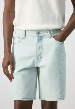 Personne portant un short en jean bleu clair et un haut blanc sans manches, une main dans la poche et l'autre détendue à côté.