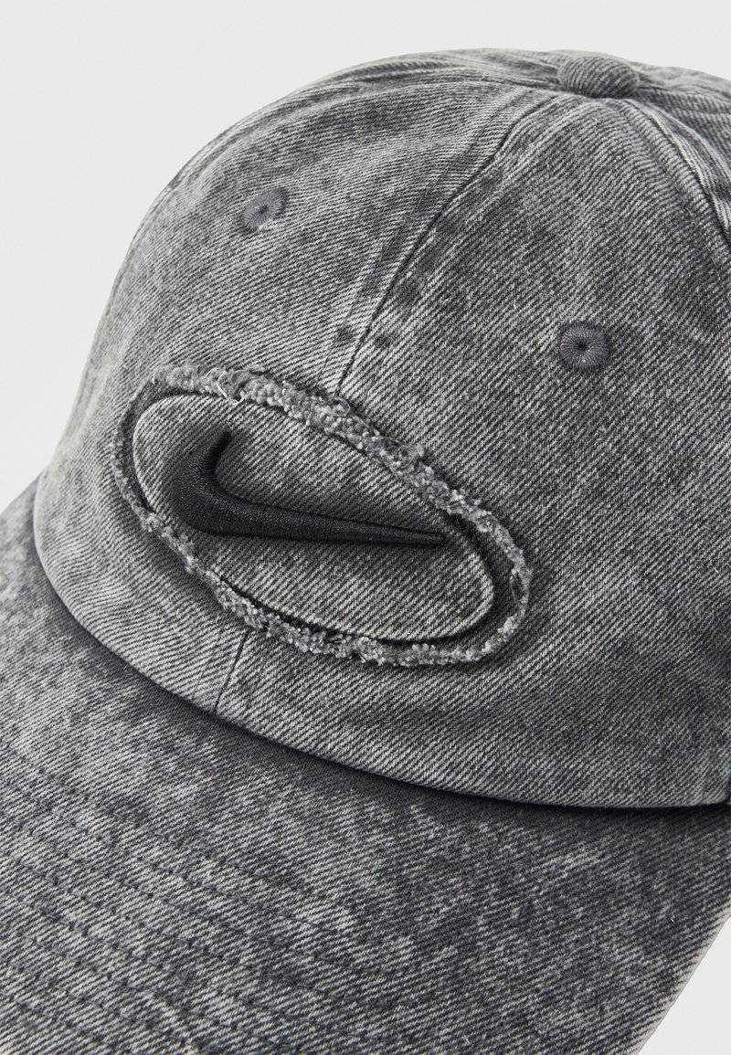 Casquette de baseball en denim gris avec un logo swoosh noir brodé, entouré d'un patch ovale effiloché sur le panneau avant.