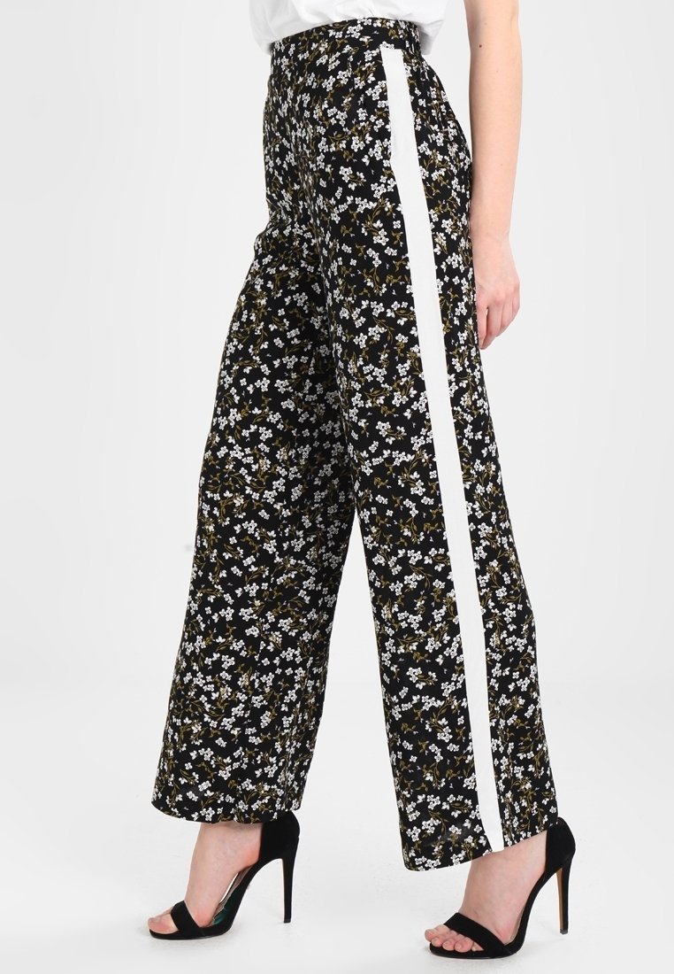 Pantalons larges à motifs floraux noirs avec des rayures blanches sur les côtés, présentant une taille haute et un tissu fluide. Accentué de petites fleurs blanches.