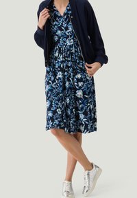 Vestido floral azul marino con patrones en azul y blanco, combinado con un cárdigan oscuro y zapatillas plateadas. Tejido suave, diseño a la altura de la rodilla.