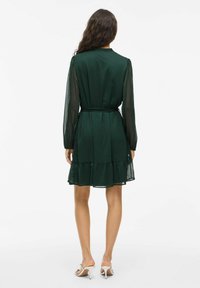 Rochie verde cu un design în straturi, mâneci lungi din material transparent și talie accentuată cu un brâu. Fabricată dintr-un material ușor, cu o textură netedă.