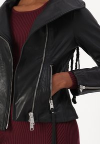 Crna kožna jakna s velikim ovratnikom, srebrnim patentnim zatvaračima i bočnim džepovima. Materijal je gladak s prilagođenim krojem. Naglasci uključuju kontrastni burgundski rebrasti pulover.