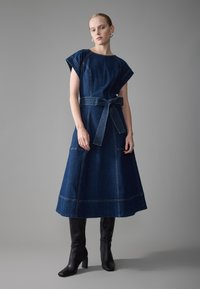 Robe midi en denim bleu foncé avec des manches courtes et une taille ceinturée. Elle présente des coutures visibles et un ourlet évasé. Associée à des bottes noires arrivant au genou.