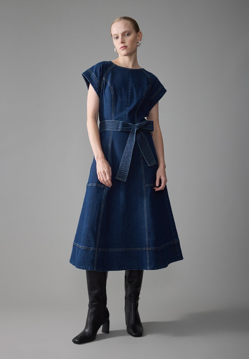 Robe midi en denim bleu foncé avec des manches courtes et une taille ceinturée. Elle présente des coutures visibles et un ourlet évasé. Associée à des bottes noires arrivant au genou.