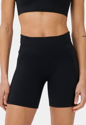 Vrouw draagt zwarte biker shorts met hoge taille en een zwart croptop, waarbij de buik en tatoeage op de linkerhand en rechteronderarm zichtbaar zijn.