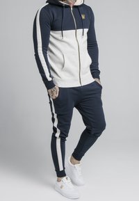 Felpa con zip blu navy e grigia chiara, con tasche, pantaloni della tuta svasati coordinati e sneakers bianche; presenta dettagli rigati e logo dorato.