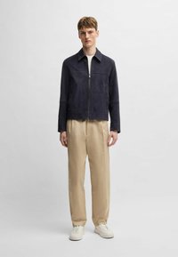 Suede navy jacket with a zippered front, pointed collar, and long sleeves, paired with beige trousers and white sneakers.