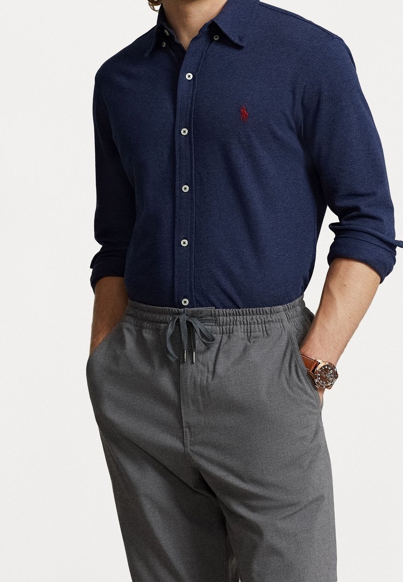 Mörkblå långärmad polo t-shirt med en röd logotyp, ihop med grå byxor med dragsko. Har knäppning fram och uppvikta ärmslut.