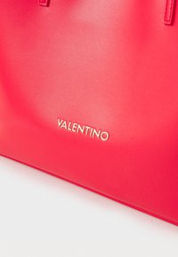 Bolsa de mão em couro vermelho com textura brilhante, apresentando o logotipo dourado "Valentino" em destaque na parte frontal. Com duas alças superiores.