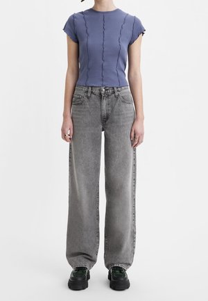 Personne portant un haut bleu ajusté à manches courtes avec des coutures ondulées, un jean gris ample et des chaussures noires à plateforme épaisse sur un fond blanc.