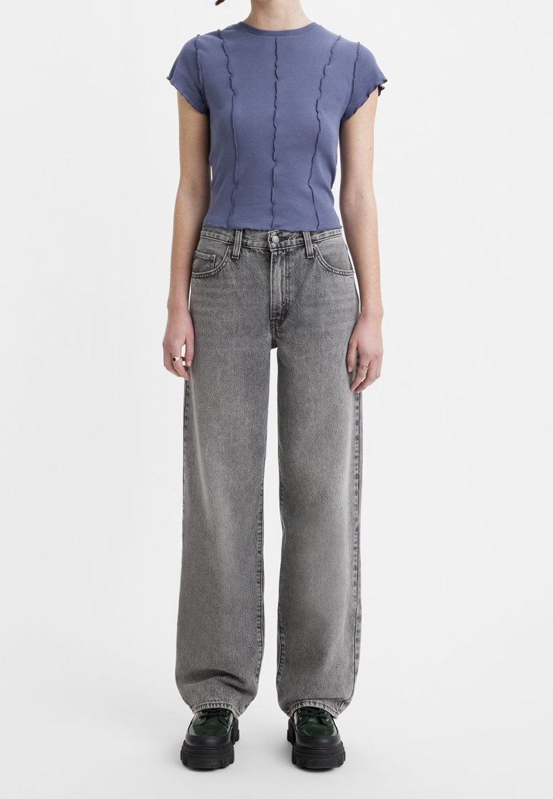 Personne portant un haut bleu ajusté à manches courtes avec des coutures ondulées, un jean gris ample et des chaussures noires à plateforme épaisse sur un fond blanc.