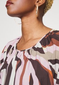 Färgglad blus med en samlad halsringning, abstrakta mönster i rosa, brunt och krämfärg, samt ett mjukt, lätt tyg. Guldiga hoop-örhängen.