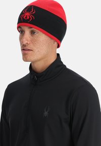 Schwarze und rote Mütze mit Spinnenlogo, gestrickte Textur und horizontales Farbblockdesign. Getragen mit einem schwarzen Quarter-Zip-Oberteil.