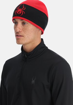 Schwarze und rote Mütze mit Spinnenlogo, gestrickte Textur und horizontales Farbblockdesign. Getragen mit einem schwarzen Quarter-Zip-Oberteil.