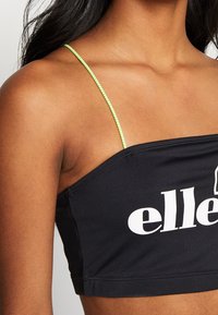 Top court noir avec un décolleté droit, doté de fines bretelles vert néon et d'un lettrage blanc audacieux à l'avant. Texture lisse.