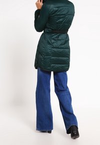 Zelená péřová bunda s opaskem v pase a texturovaným povrchem, s délka ke kolenům. Nosit s širokými modrými džíny.
