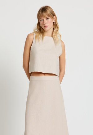 Donna bionda con capelli ondulati indossa un top beige senza maniche e una gonna a vita alta, in piedi davanti a uno sfondo bianco uniforme.