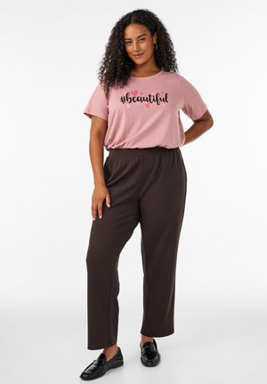 Frau mit lockigem Haar, die ein pinkes T-Shirt mit dem Text "#beautiful" und Herzen trägt, braune Hosen und schwarze Loafers, steht mit einer Hand in der Hüfte.