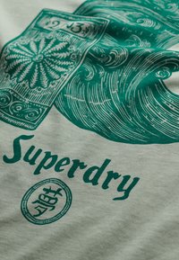 Tela de color gris claro con diseño impreso en verde, que incluye olas en espiral y elementos decorativos. El texto dice "Superdry" con un embellecimiento circular.