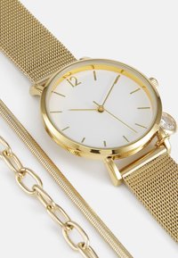 Montre avec bracelet en maille dorée, cadran blanc, index horaires dorés, un petit accent en cristal et un design minimaliste. Comprend une chaîne dorée et un maillon texturé.