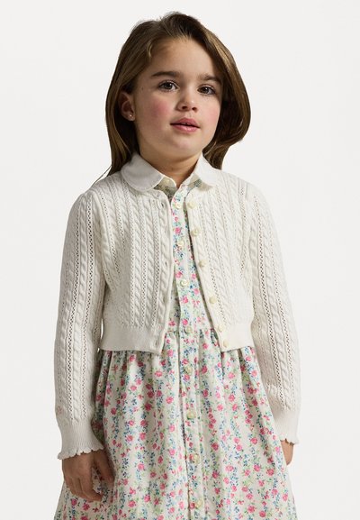 White knitted cardigan with cable patterns, round neckline, and button closure, worn over a colorful floral print dress with short sleeves.